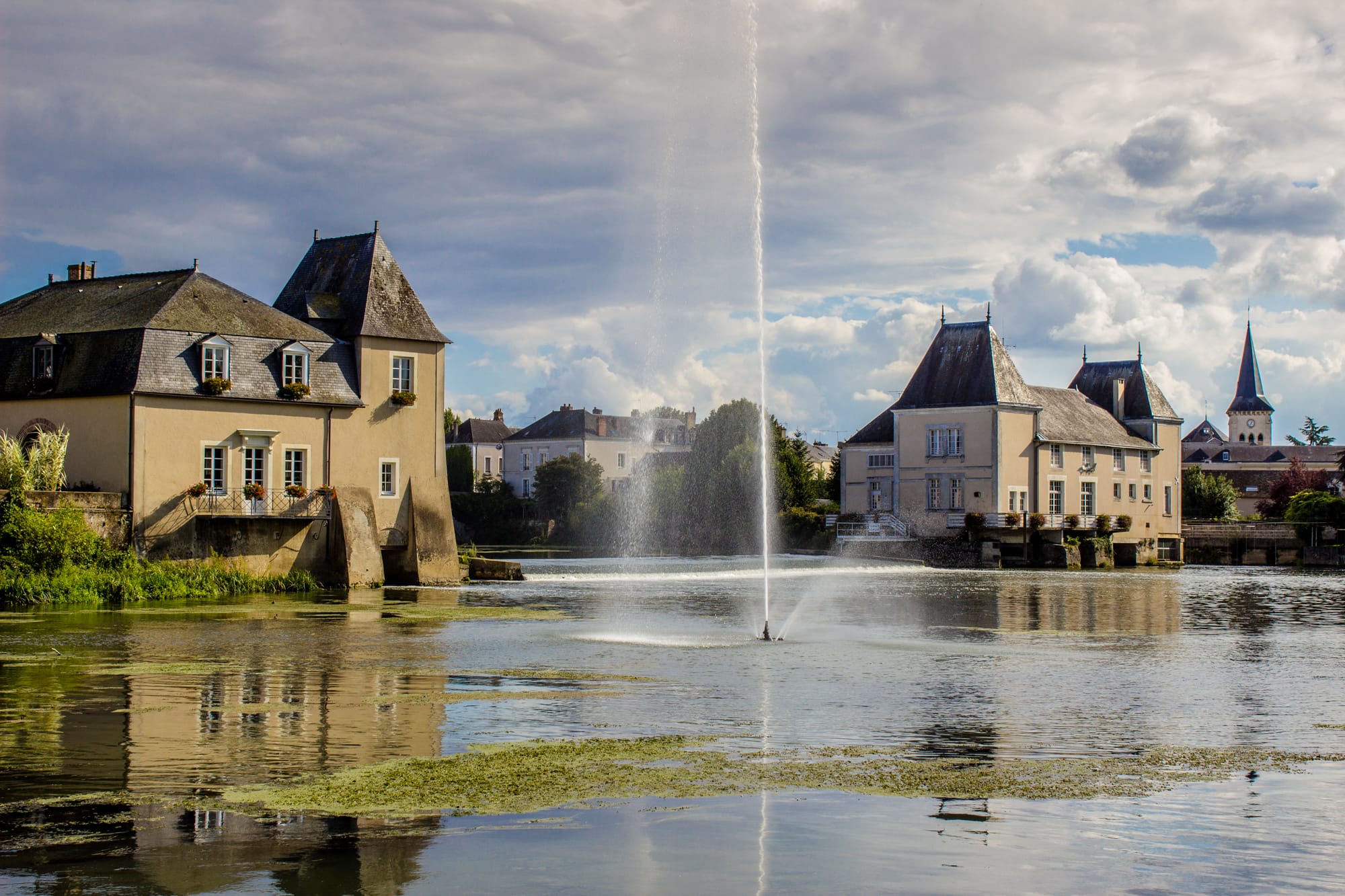 Vue pittoresque du château de La Flèche en bord de Loir, avec ses bâtiments historiques reflétés dans l’eau et une fontaine centrale animant l’étang