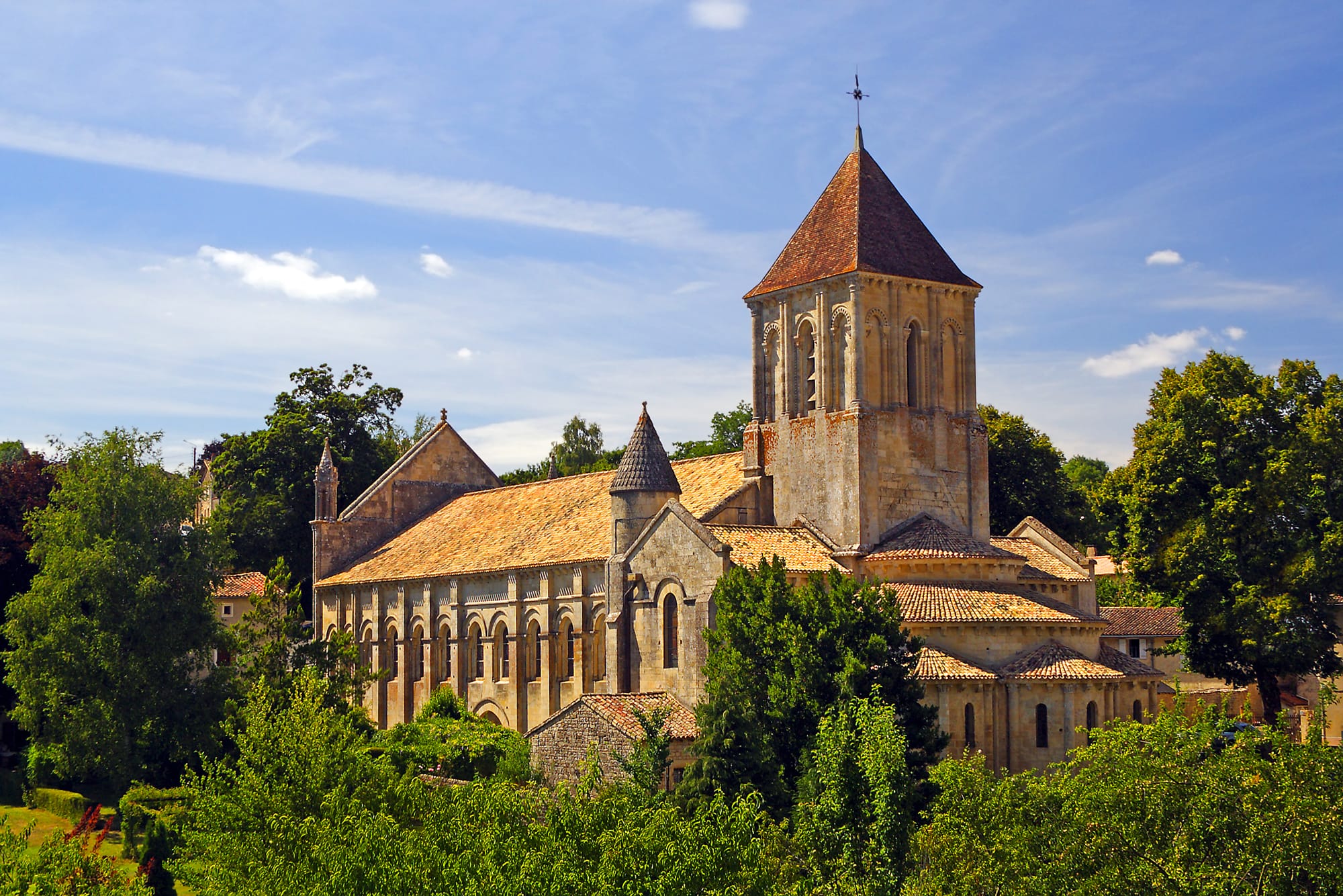 Église romane Saint-Hilaire de Melle avec son clocher imposant et ses toits de tuiles, entourée de verdure et baignée de soleil
