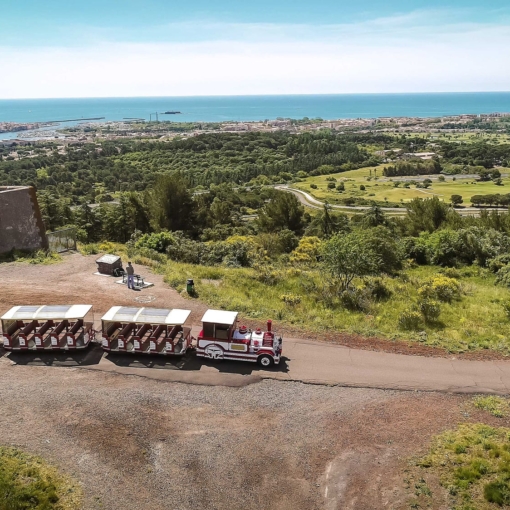 Petit train touristique blanc et rouge circulant sur une route en hauteur avec vue sur la mer et la ville du Cap d’Agde.