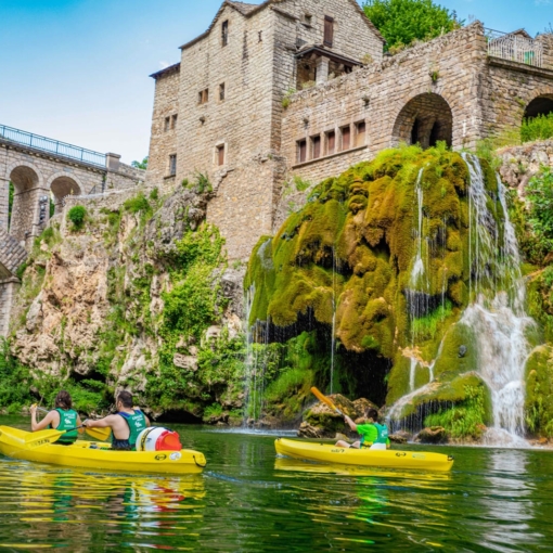 Canoë sur la rivière du Tarn passant sous un pont de pierre et près d’une cascade verdoyante dans les Gorges du Tarn