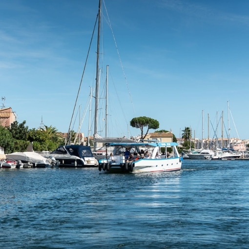 Bateau à passagers naviguant sur un canal bordé de voiliers et de maisons à Agde sous un ciel ensoleillé.