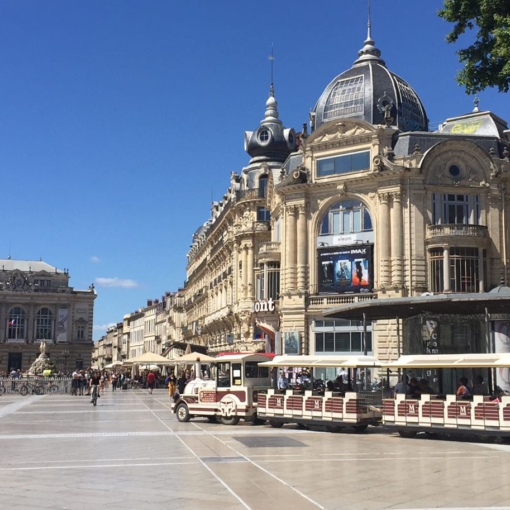 Petit train touristique circulant sur la place de la Comédie à Montpellier devant les façades haussmanniennes et l’Opéra.